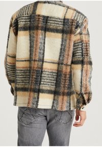 Wełniana koszula w kratę w odcieniach beżu, czerni i pomarańczu. Posiada luźny krój, mankiety na guziki oraz boczne rozporki dla lepszego ruchu.