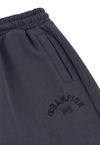 Pantalons de jogging gris foncé avec une taille élastique, dotés d'une poche latérale et d'un logo "Champion" imprimé en noir. Texture lisse et design décontracté.