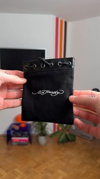 Schwarze Canvas-Tasche mit einem glänzenden schwarzen oberen Rand, Metallösen und einem Kordelzugverschluss. Mit weiß gesticktem Schriftzug "Ed Hardy".