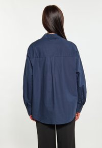 Námořnická modrá, oversized košile na knoflíky vyrobená z bavlny s klasickým límcem, záhybovým detailem na zadní straně a dlouhými, objemnými rukávy.