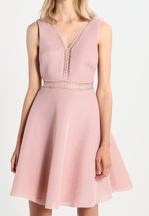 Robe rose sans manches avec un col en V, des détails en crochet à l'encolure et à la taille, et une jupe évasée portée par une personne debout.