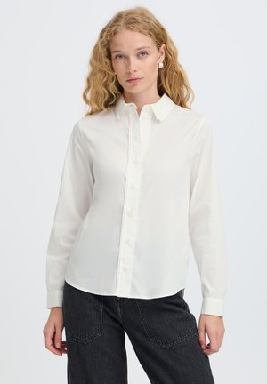 Femme aux cheveux blonds bouclés portant une chemise blanche boutonnée et un jean foncé sur un fond clair uni.