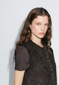 Junge Frau mit langen, welligen braunen Haaren, die ein schwarzes T-Shirt mit kurzen Ärmeln und eine geknöpfte, dunkel strukturierte Weste trägt. Sie blickt nach vorne mit einem neutralen Ausdruck.