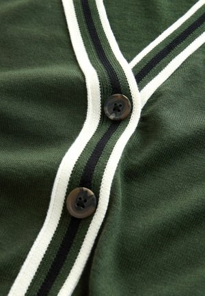 Grüne Strickjacke mit weißem und schwarzem gestreiftem Blendenbesatz und zwei dunklen Knöpfen. Der Stoff hat eine glatte Textur.