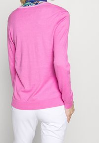 Růžový bavlněný svetr s dlouhými rukávy a žebrovanými manžetami, s kulatým výstřihem. Nositelný přes bílé kalhoty. Hladká textura, lehký design.