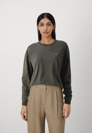 Chemise en coton à manches longues, de couleur verte foncée, avec un col rond et un petit logo imprimé. Assortie à un pantalon taille haute beige.