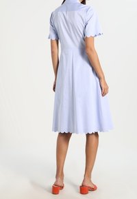 Ljusblå klänning med en åtsittande bodice och en utställd kjol. Har scalloped ärmslut och en slät textur. Bärs med orange sandaler.