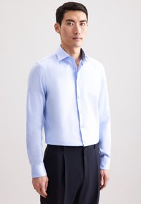 Man gekleed in een lichtblauw overhemd met knopen en zwarte broeken, staande tegen een effen witte achtergrond, recht naar voren kijkend met een neutrale uitstraling.