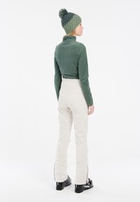 Maglione verde in pile a maniche lunghe, pantaloni beige a vita alta con cerniere alle caviglie e un cappello lavorato a maglia multicolore con pom-pom.