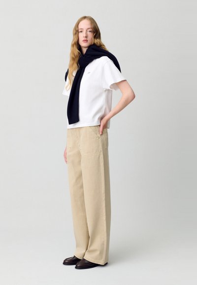 Camiseta de algodón blanca, de corte suelto, combinada con pantalones de pierna ancha beige con sutiles rayas. Suéter azul marino oscuro atado a los hombros. Zapatos marrones.