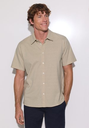 Mann mit lockigem Haar, der ein beiges kurzärmeliges Hemd mit kleinem geometrischem Muster und dunkle Hose trägt, lächelt und schaut seitwärts.