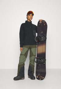 Pullover in pile nero con dettagli a bottoni, pantaloni da snowboard verde oliva con toppe, snowboard nero con attacchi e occhialini arancioni.