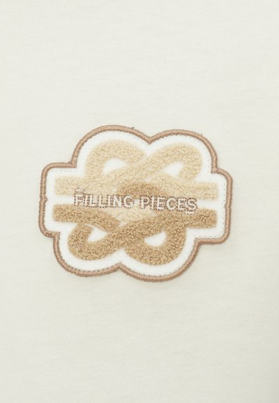 Filling Pieces izšūts logo bēšā un baltā krāsā uz gaiša auduma fona, kas iekļauj teksturētu, mākonis līdzīgu dizainu, ko ieskauj brūna kontūra.