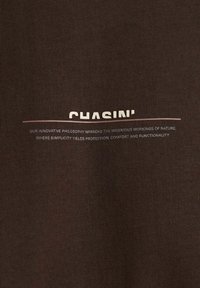 Bruine stof met de tekst "CHASIN'" boven een dunne horizontale lijn en daaronder kleinere tekst die over filosofie, natuur, bescherming, comfort en functionaliteit gaat.