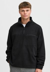 Pullover in pile nero con colletto alto, chiusura a mezza zip e tasca frontale. Presenta una superficie testurizzata ed è di vestibilità ampia.