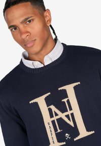 Suéter de punto navy con un gran logotipo en crema de "HN" y "HARPER" debajo. El cuello muestra una camisa de vestir a rayas en azul claro y blanco.