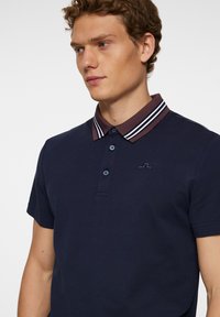Navyblå polo-shirt med korta ärmar, med en krage i brun och vit rand, två-knapps knappslå och en diskret logotyp på bröstet.