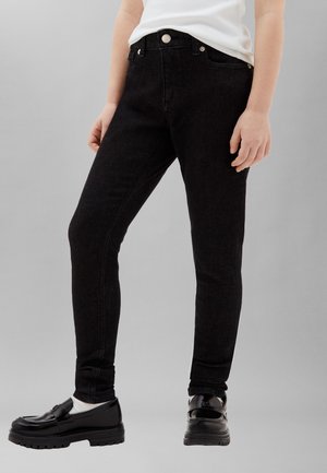 Isik, kes kannab musti slim-fit teksapükse, valgeid sokke, musti loafer-tüüpi jalanõusid ja valget täringut hallil taustal.
