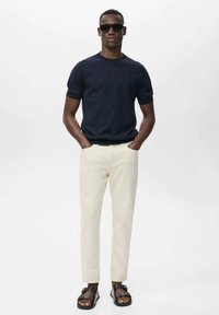 Tricou tricotat de culoare bleumarin cu mânecă scurtă, pantaloni slim-fit bej și sandale negre. Modelul stă cu mâinile în buzunare, pe un fundal alb.