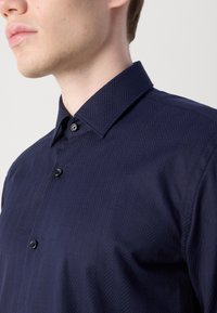 Navy blue shirt featuring a herringbone pattern, button-down collar, and a smooth texture. Black buttons accent the front placket.