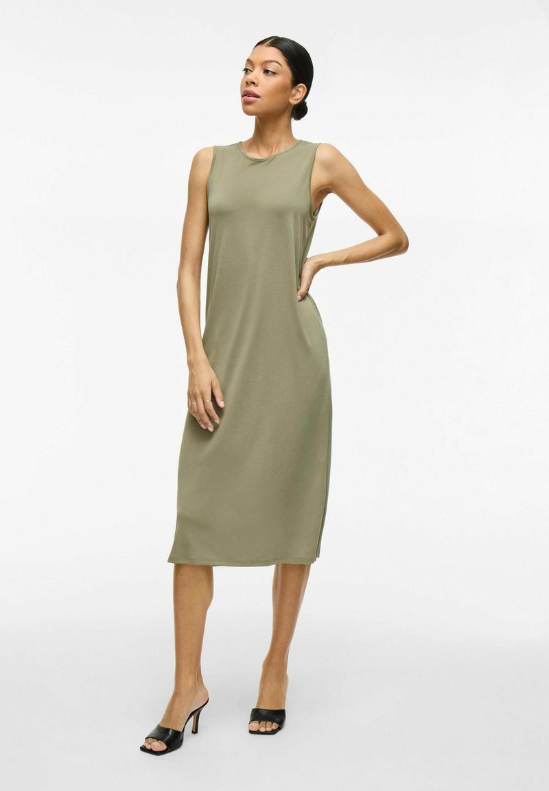 Oliwkowa, bezrękawnikowa sukienka midi wykonana z miękkiego materiału, charakteryzująca się prostym wzorem i bocznymi rozcięciami. Połączona z czarnymi, odkryto-palcowymi szpilkami.