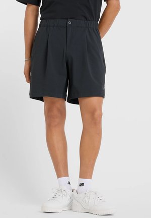 Uomo che indossa pantaloncini neri con vita elastica, calzini bianchi New Balance alla caviglia e sneakers bianche New Balance, in piedi contro uno sfondo uniforme.