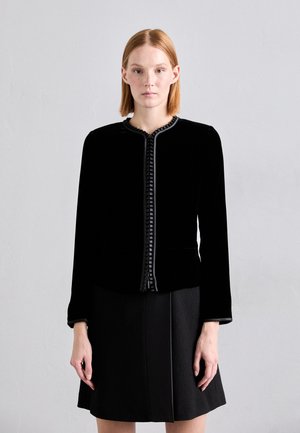 Black velvet jacket with a round neckline, decorative ribbed trim, long sleeves, and front pockets, paired with a textured black skirt.