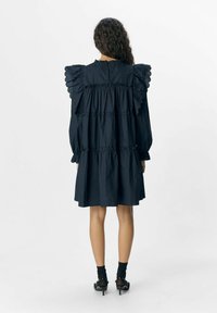 Schwarzes Kleid mit Rüschensäumen, strukturiertem Stoff, stufenweise Gestaltung und lässiger Passform. Mit Knöchelstrümpfen und Absatzschuhen gestylt.