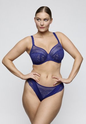 Vrouw poseert in bijpassende blauwe kanten beha en slip met handen op de heupen tegen een effen witte achtergrond.