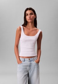 Débardeur blanc ajusté avec un décolleté rond et un détail de logo subtil au centre, associé à un jean taille haute bleu clair.
