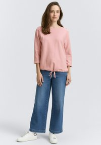 Helles Rosa, langärmliges Oberteil mit rundem Halsausschnitt undKordelzugsaum, kombiniert mit weiten blauen Jeanshosen und weißen Turnschuhen.