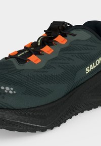 Σκούρο πράσινο και μαύρο αθλητικό παπούτσι Salomon με πορτοκαλί θηλιές κορδονιών και γρήγορο σύστημα δέσιμου σε λευκό φόντο.