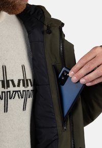 Tmavě zelená bunda se zipovou kapsou, s černou podšívkou. Do kapsy se vkládá modrý chytrý telefon. Viditelný texturovaný svetr.