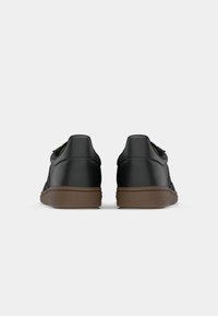 Sneakers sintetici neri con superficie texturizzata, tacco arrotondato e suola in gomma marrone, visti da dietro. Design semplice, senza loghi visibili.