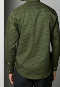 Green patterned long-sleeve shirt featuring a geometric design, button cuffs, and a curved hem, made from a smooth, lightweight fabric.