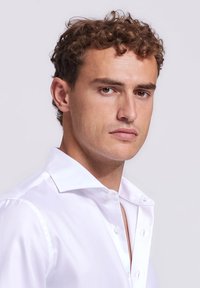 Witte overhemd met een gestructureerde kraag, knoopsluiting aan de voorkant en een gladde afwerking, gedragen door een model met kort, krullend haar.