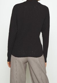 Czarny sweter z prążkowanego dzianiny z okrągłym dekoltem, dopasowany kształt i teksturowana tkanina, w połączeniu z beżowymi spodniami w czarne kratki.