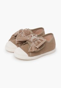 Kleine beigefarbene Wildlederschuhe für Kleinkinder mit abgerundeten weißen Gummisohlen und großen dekorativen Samtbinden oben, auf einem weißen Hintergrund präsentiert.