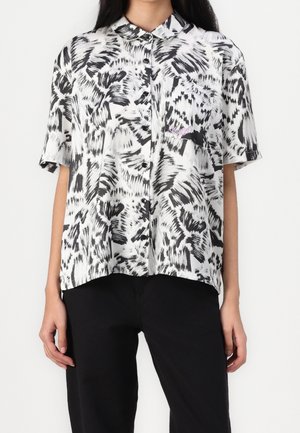 Femme portant une chemise à manches courtes boutonnée avec un motif abstrait en coups de pinceau noir et blanc, et un pantalon noir.