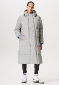 Vrouw in een lange lichtgrijze gewatteerde winterjas met capuchon, zwarte leggings, witte sokken met zwarte strepen en witte sneakers.