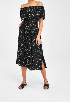 Frau in einem schwarzen, schulterfreien Midi-Kleid mit weißen Punkten, seitlichem Schlitz und schwarzen flachen Sandalen, steht mit den Händen in den Taschen.