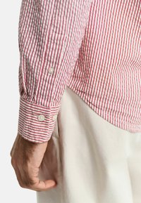 Rood en wit gestreepte knoopshirt met lange mouwen, met een ronde zoom en witte knopen. De stof lijkt textuur te hebben en is lichtgewicht.