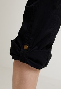 Zwarte broek met opgerolde boorden heeft een gladde textuur, een knoopaccent op de boorden en een taps toelopende pasvorm bij de enkels.