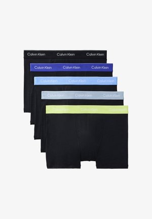 Oben ohne stehender Mann, barfuß, trägt schwarze Calvin Klein Boxershorts und richtet mit der Hand den Bund, vor schlichtem grauem Hintergrund.