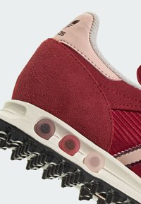 Κόκκινο αθλητικό παπούτσι από σουέντ και ύφασμα με λεπτομέρεια από ροζ δέρμα, διαθέτοντας υφασμένη μαύρη ελαστική σόλα και τρεις πολύχρωμες κυκλικές λεπτομέρειες.