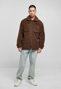 Brun fleecejakke med krave, to brystlommer og knaplukning. Båret over lyseblå jeans og hvide sneakers. Blød tekstur.