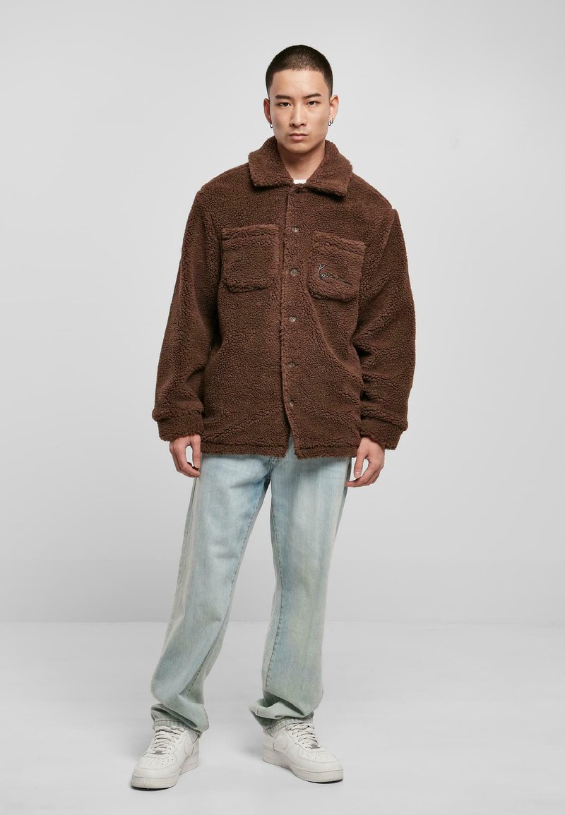 Brun fleecejakke med krave, to brystlommer og knaplukning. Båret over lyseblå jeans og hvide sneakers. Blød tekstur.