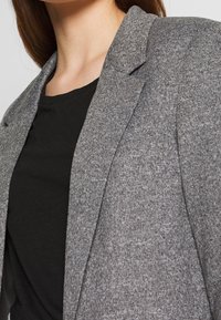 Szara marynarka z teksturowanej tkaniny, z kołnierzem z wycięciem i otwartym przodem. Nosi się ją na czarnej koszuli, prezentując styl warstwowy.