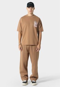 Kahvan puuvillat-paita taskugrafiikalla, yhdistettynä yhteensopiviin leveälahkeisiin housuihin. Lyhyet hihat ja rento istuvuus. Musta lippis logolla.