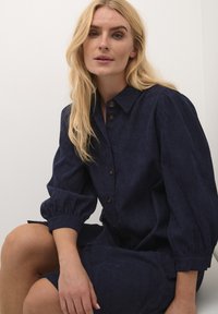 Robe chemise marine avec un col, fermeture à boutons et manches bouffantes. Fabriquée en tissu texturé, elle est de longueur genoux avec une coupe décontractée.
